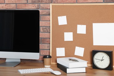 Photo of Cork board with blank paper notes, computer, books and alarm clock on wooden table near brick wall