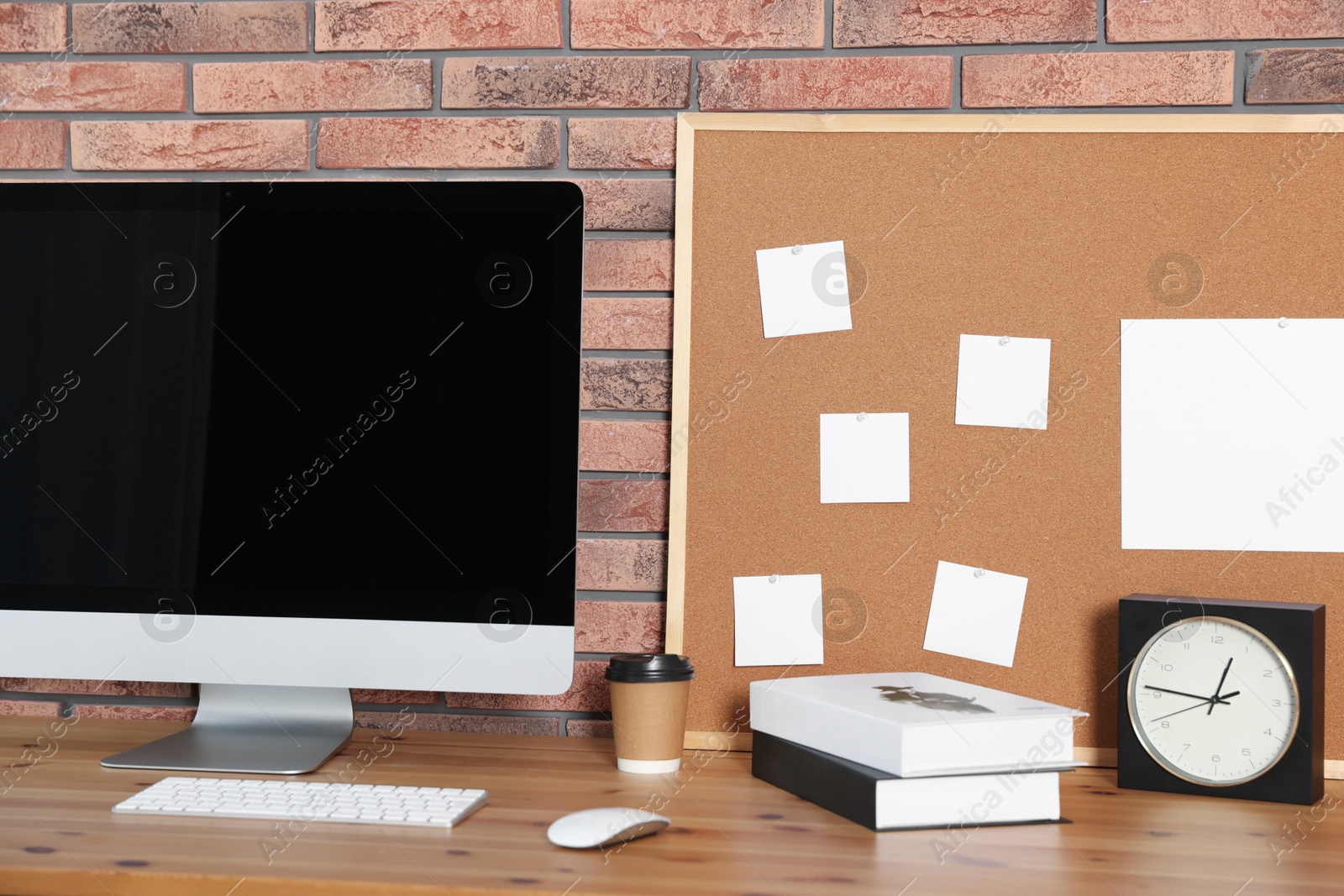 Photo of Cork board with blank paper notes, computer, books and alarm clock on wooden table near brick wall