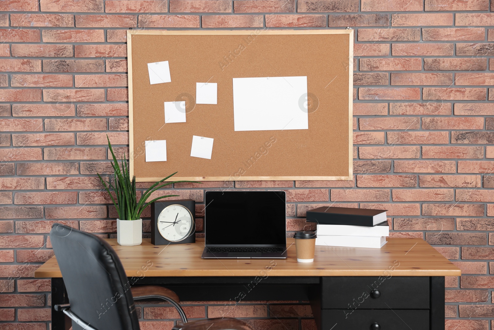 Photo of Cork board with blank paper notes, laptop, books, alarm clock and houseplant on wooden table near brick wall