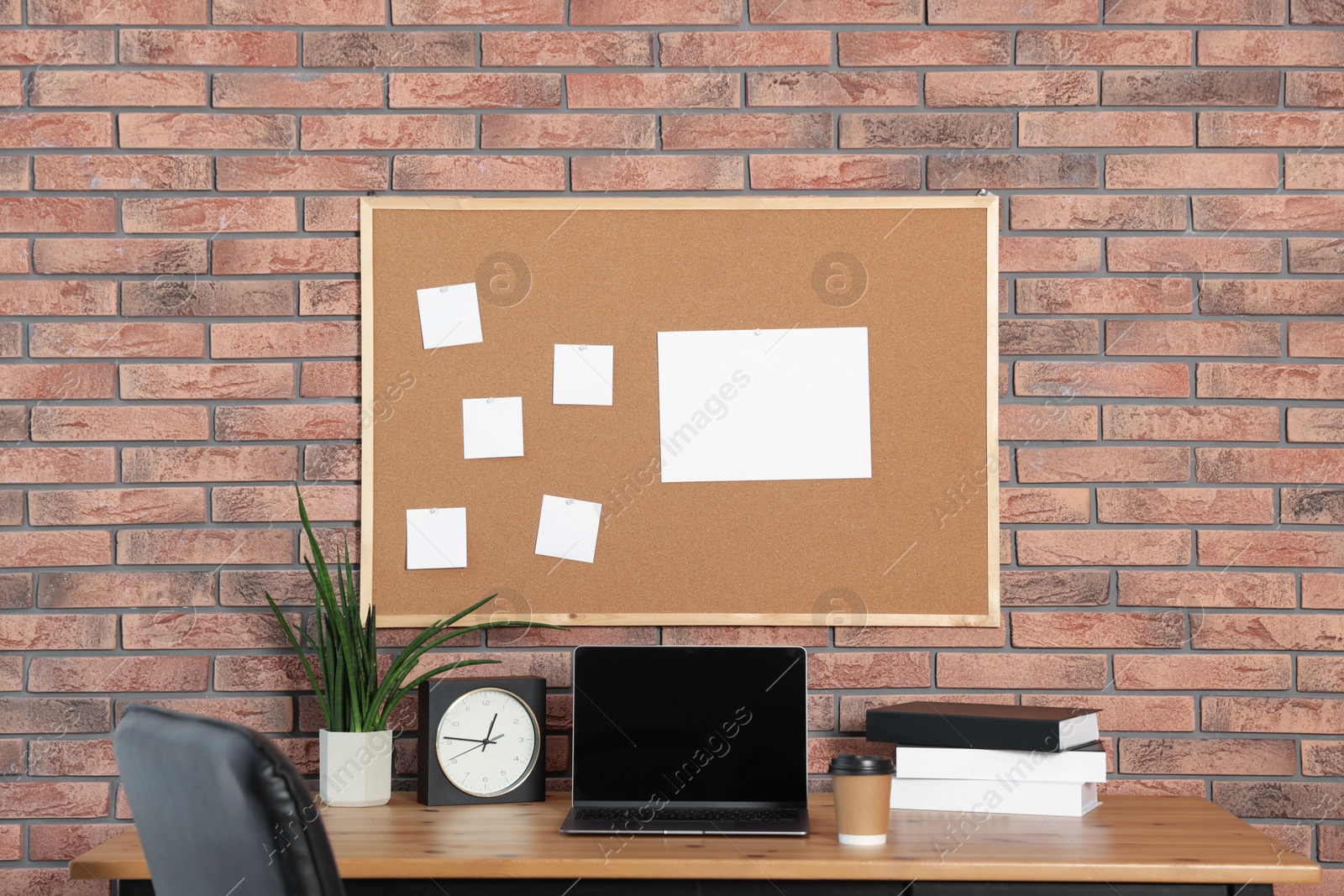 Photo of Cork board with blank paper notes, laptop, books, alarm clock and houseplant on wooden table near brick wall