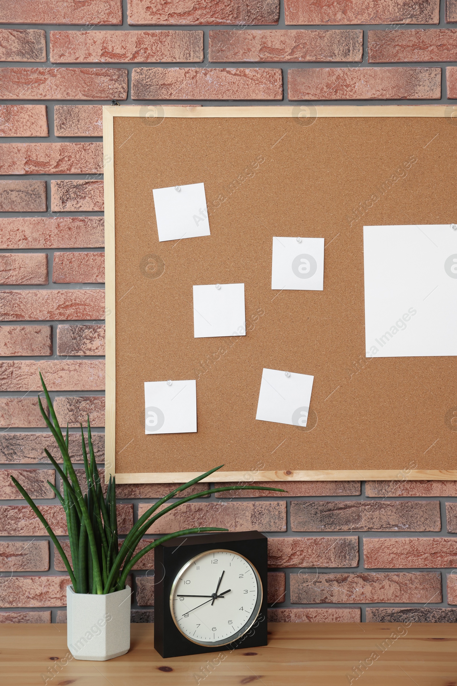 Photo of Cork board with blank paper notes, alarm clock and houseplant on wooden table near brick wall