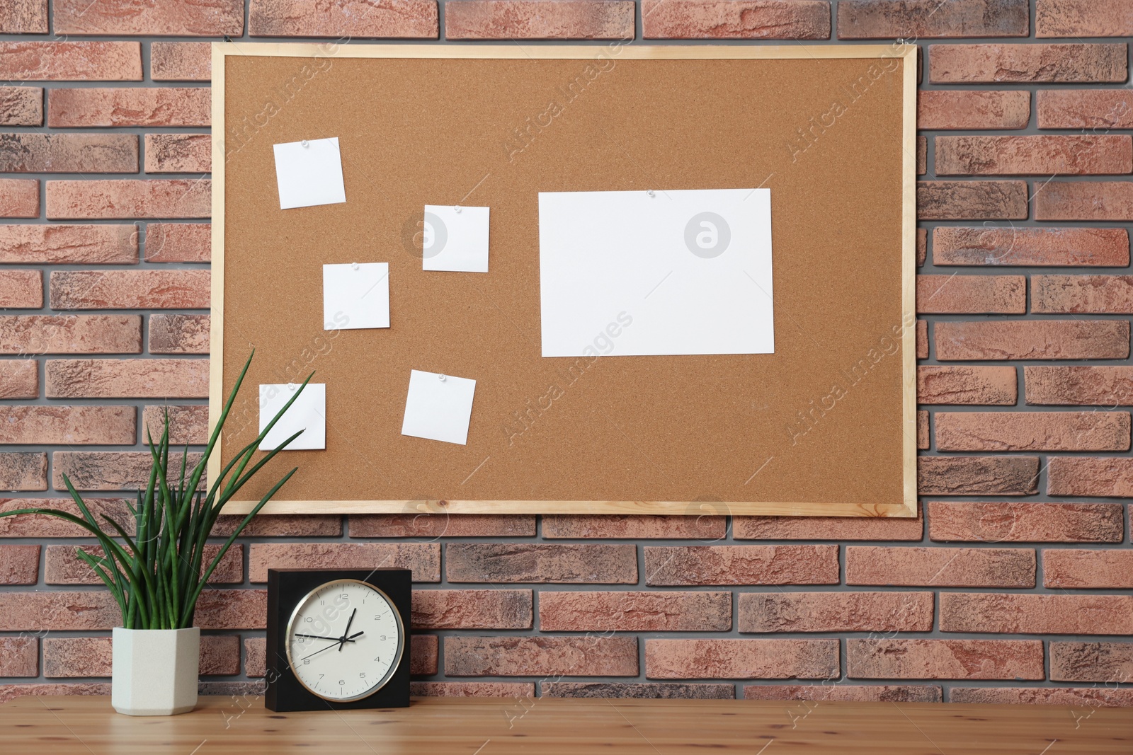 Photo of Cork board with blank paper notes, alarm clock and houseplant on wooden table near brick wall