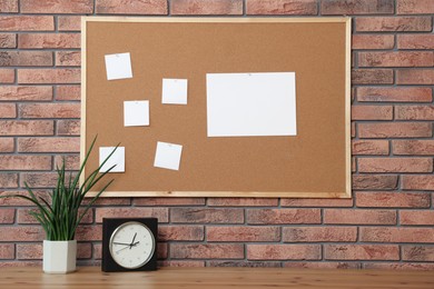 Cork board with blank paper notes, alarm clock and houseplant on wooden table near brick wall