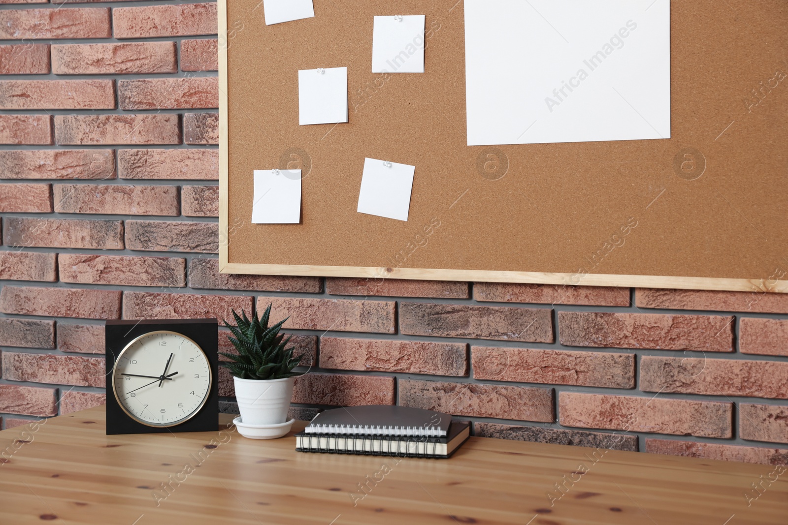 Photo of Cork board with blank paper notes, alarm clock, notebooks and houseplant on wooden table near brick wall