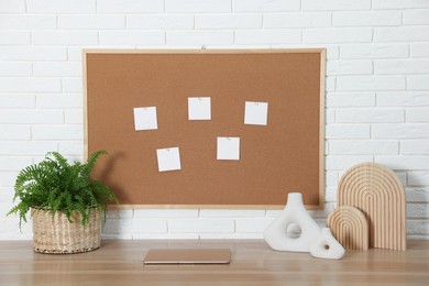 Photo of Cork board with blank paper notes, laptop and decorative elements on wooden table near white brick wall