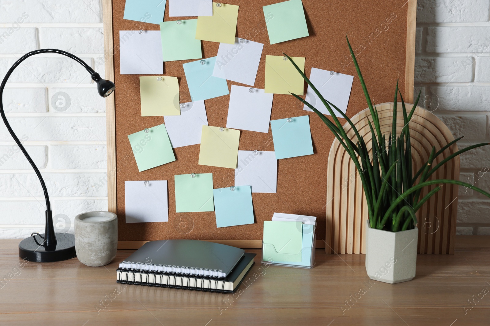 Photo of Cork board with blank paper notes, notebooks and decorative elements on wooden table near white brick wall