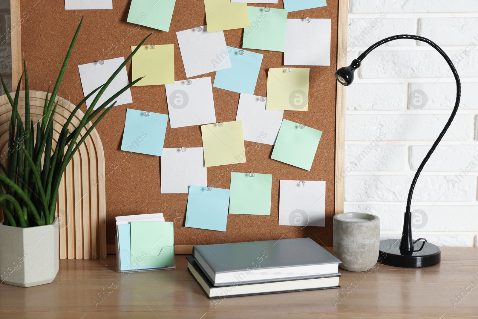 Photo of Cork board with blank paper notes, notebooks and decorative elements on wooden table near white brick wall