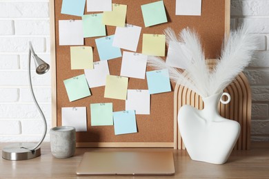 Photo of Cork board with blank paper notes, laptop and decorative elements on wooden table near white brick wall