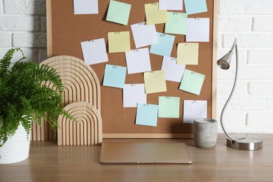 Photo of Cork board with blank paper notes, laptop and decorative elements on wooden table near white brick wall