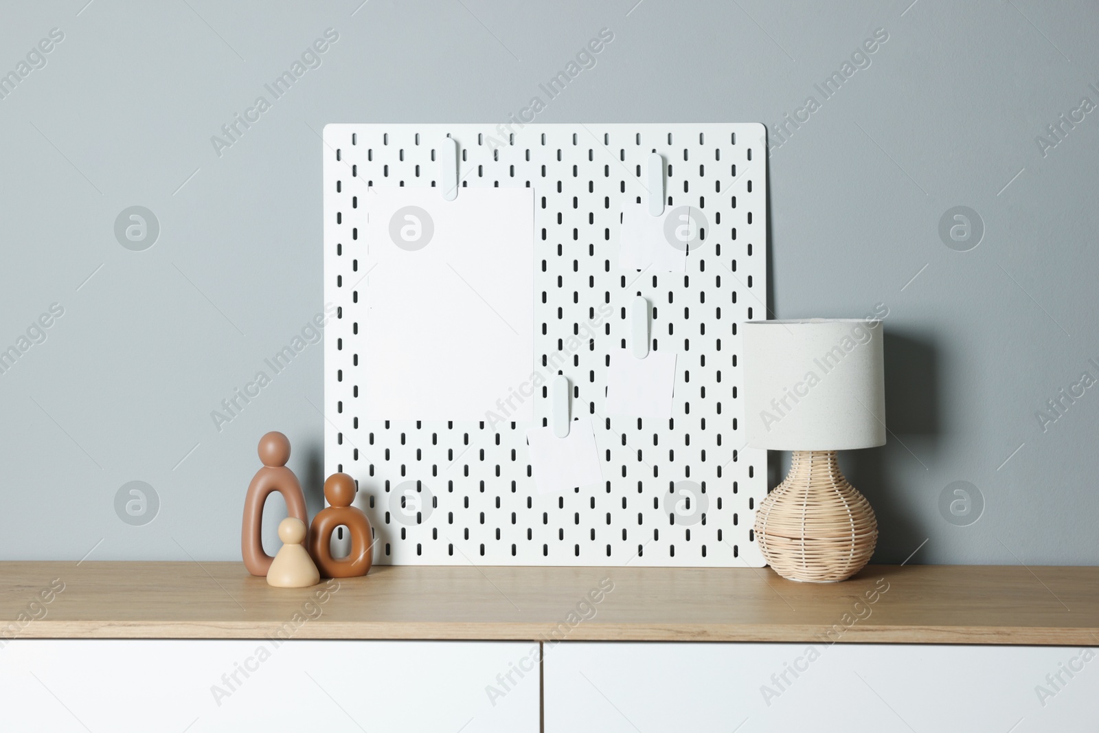 Photo of Peg board with blank paper notes, lamp and decorative figures on chest of drawers near gray wall