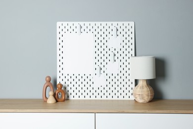 Photo of Peg board with blank paper notes, lamp and decorative figures on chest of drawers near gray wall