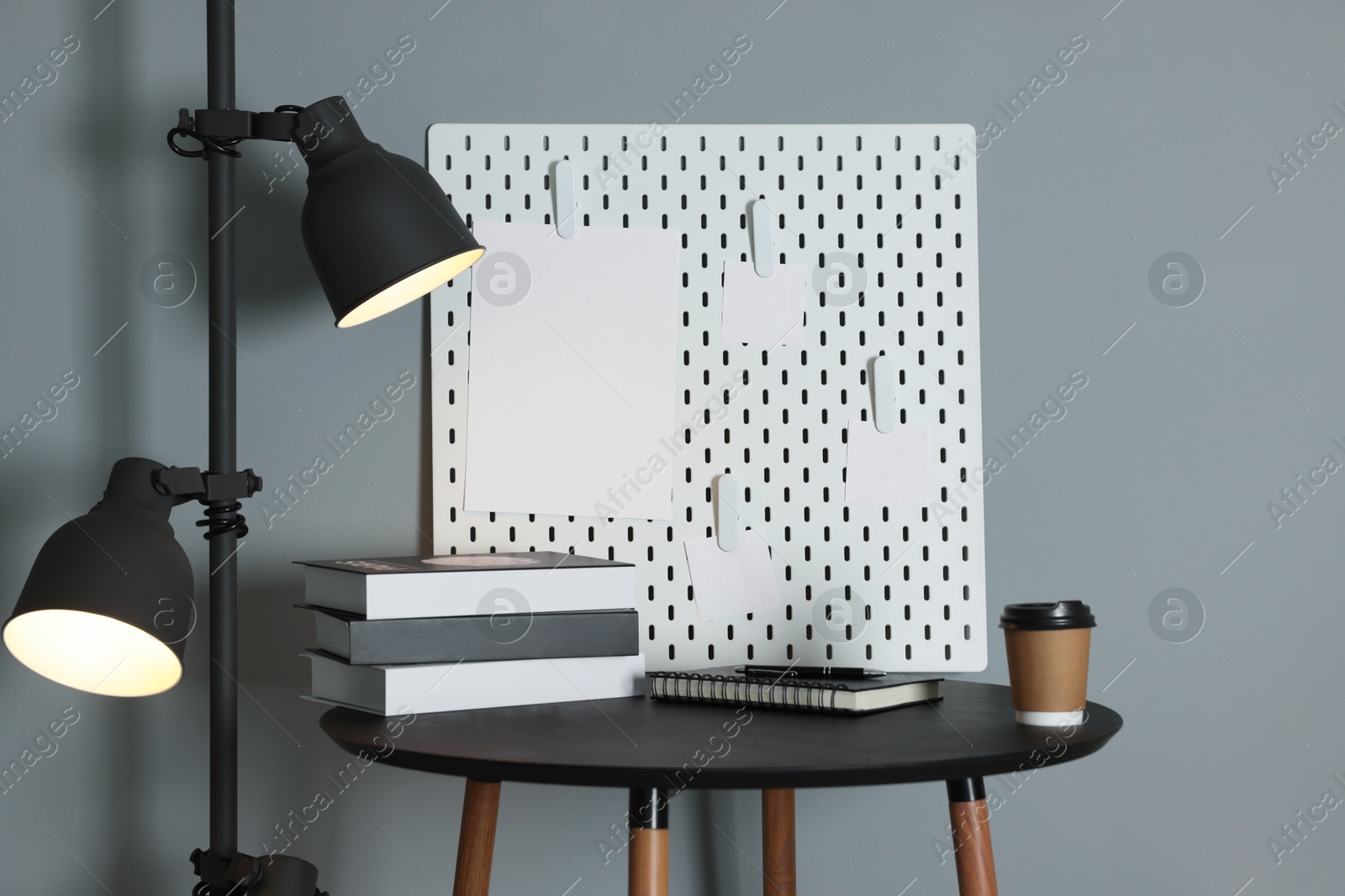 Photo of Peg board with blank paper notes, lamp, books, notebook and cup on black table near gray wall