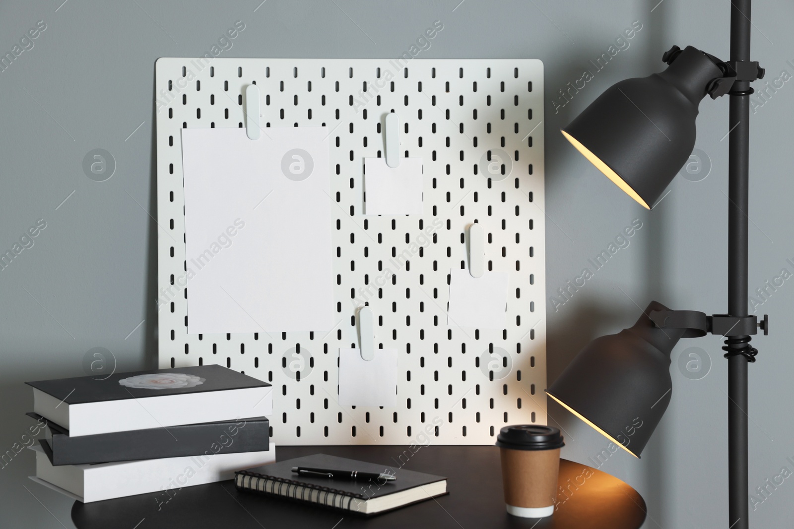 Photo of Peg board with blank paper notes, lamp, books, notebook and cup on black table near gray wall