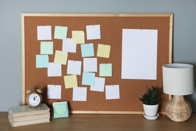 Photo of Cork board with blank paper notes, books and decorative elements on wooden table near gray wall