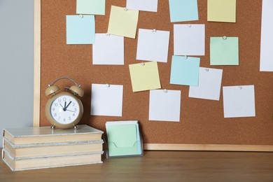 Photo of Cork board with blank paper notes, books and alarm clock on wooden table near gray wall