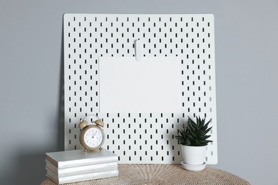 Peg board with blank paper note, books, houseplant and alarm clock on wooden table near gray wall