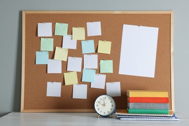 Photo of Cork board with blank paper notes, books, notebooks and alarm clock on wooden table near gray wall