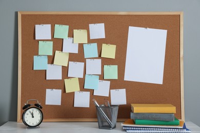 Cork board with blank paper notes, books, notebooks, pencils in holder and alarm clock on wooden table near gray wall