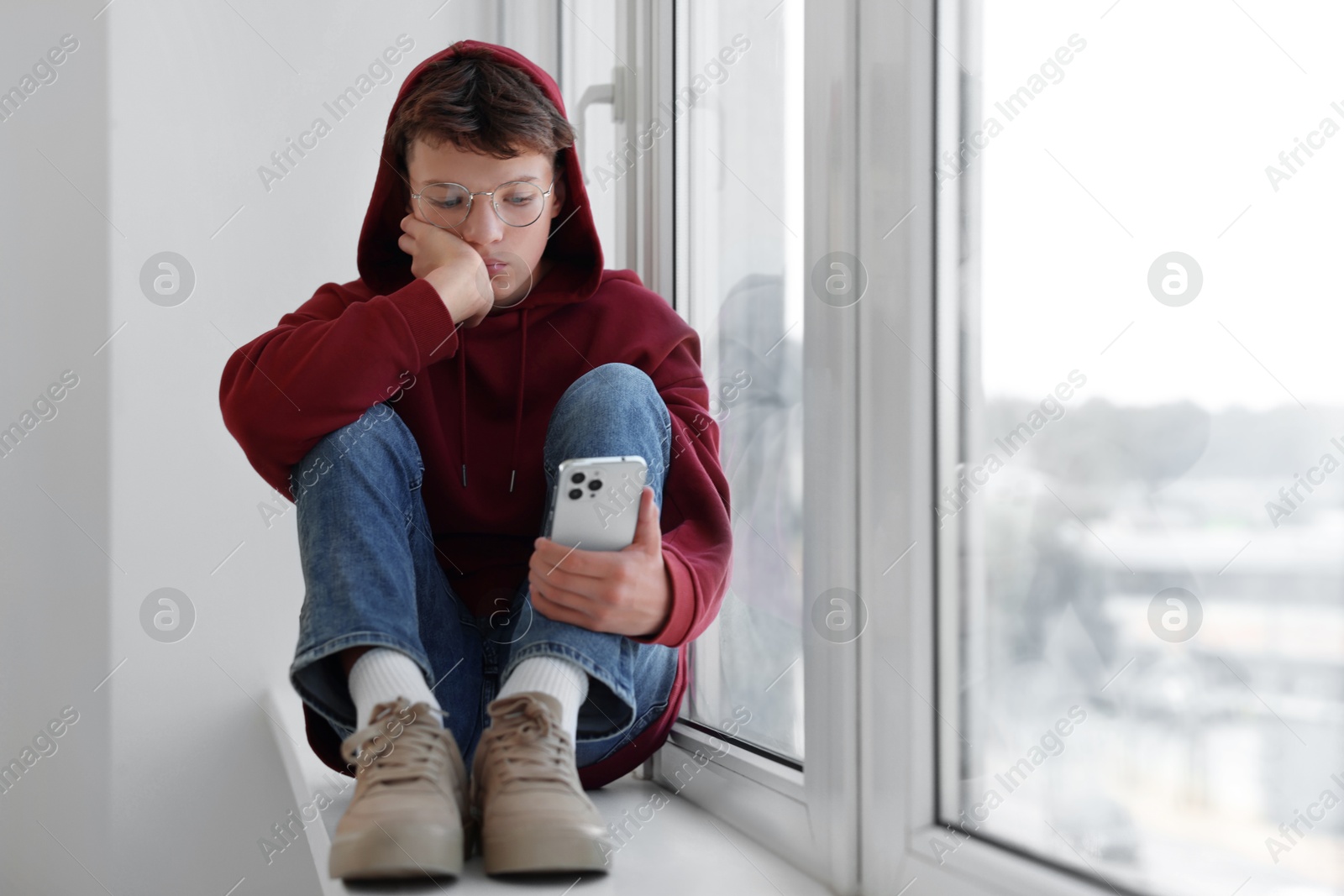 Photo of Loneliness concept. Sad teenage boy with smartphone on windowsill indoors