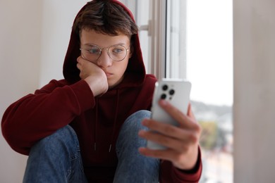 Photo of Loneliness concept. Sad teenage boy using smartphone near window indoors