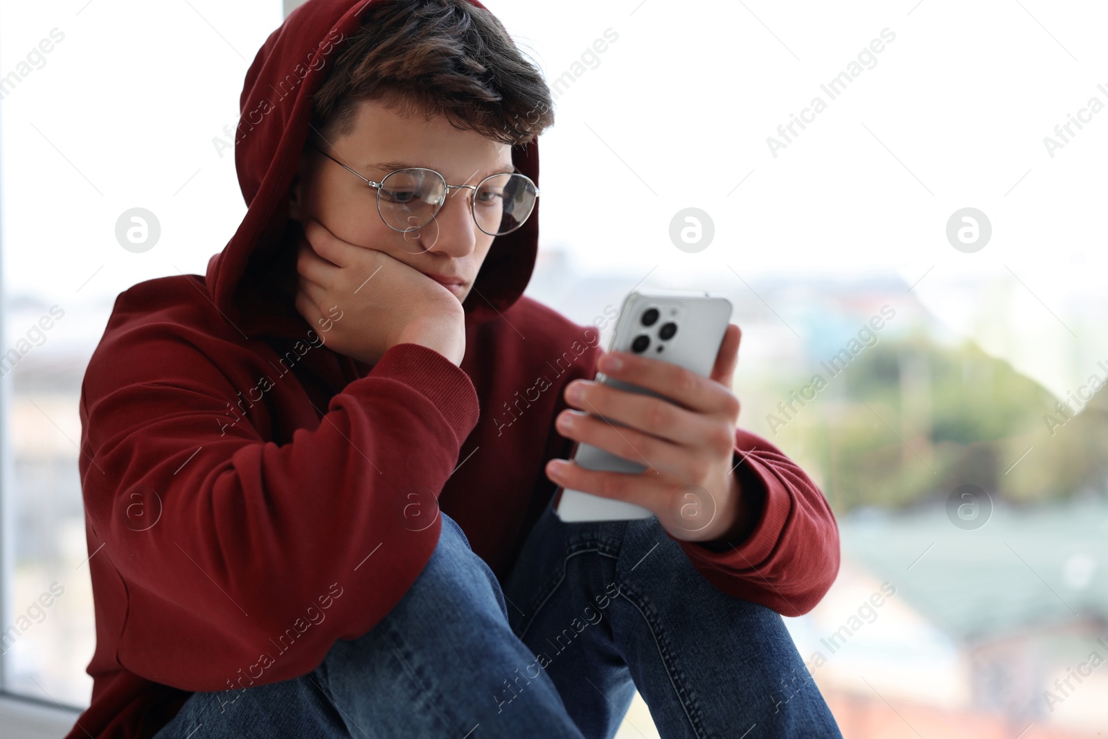 Photo of Loneliness concept. Sad teenage boy using smartphone near window indoors