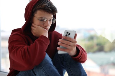 Loneliness concept. Sad teenage boy using smartphone near window indoors