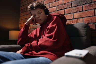 Loneliness concept. Sad teenage boy in headphones listening to music on armchair indoors