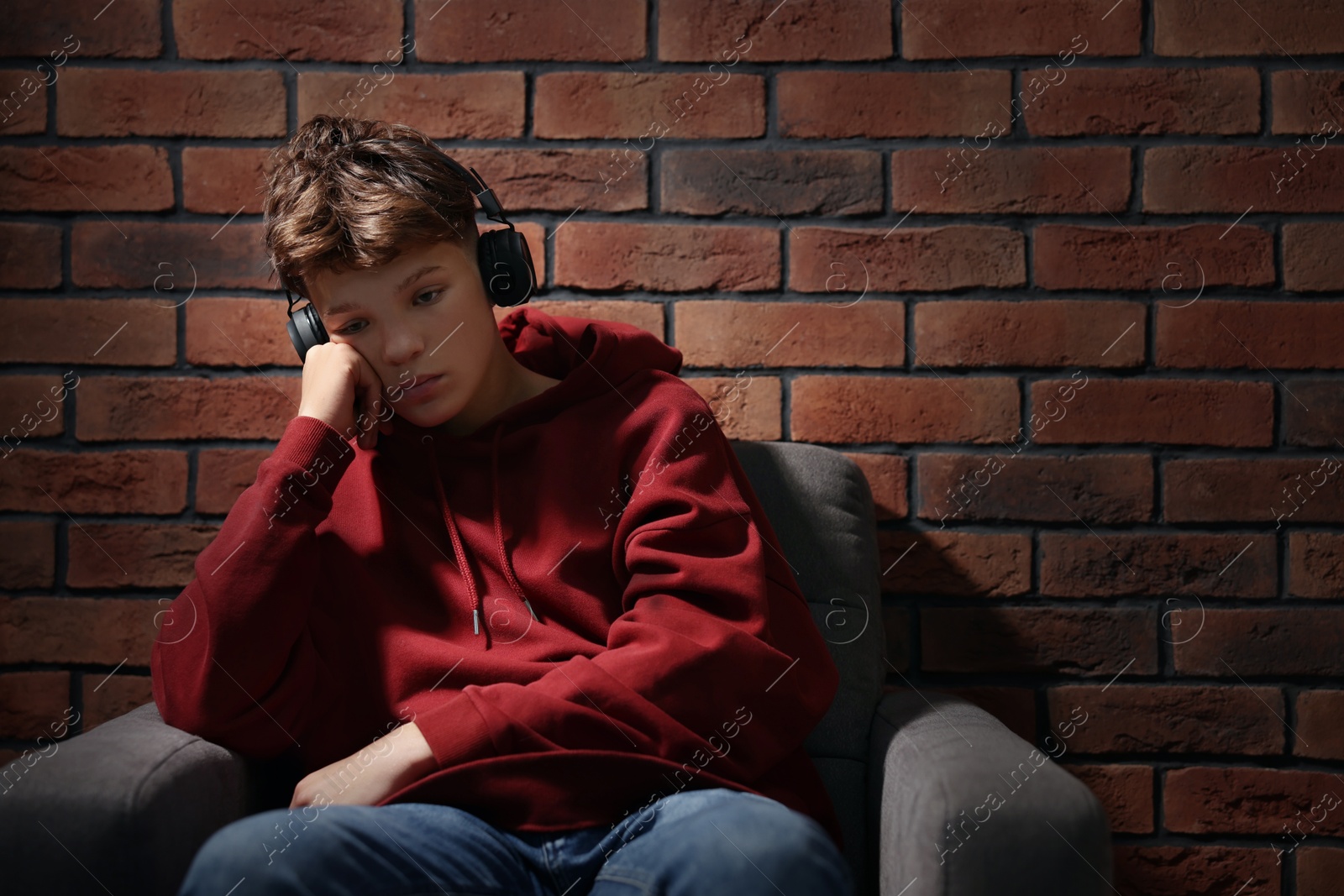 Photo of Loneliness concept. Sad teenage boy in headphones listening to music on armchair near brick wall