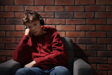 Photo of Loneliness concept. Sad teenage boy in headphones listening to music on armchair near brick wall