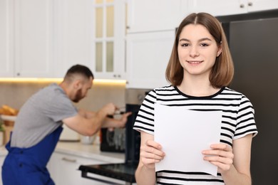 Smiling woman with sheet of paper and repairman fixing oven in kitchen, selective focus