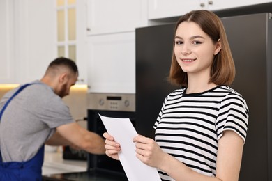 Photo of Smiling woman with sheet of paper and repairman fixing oven in kitchen, selective focus