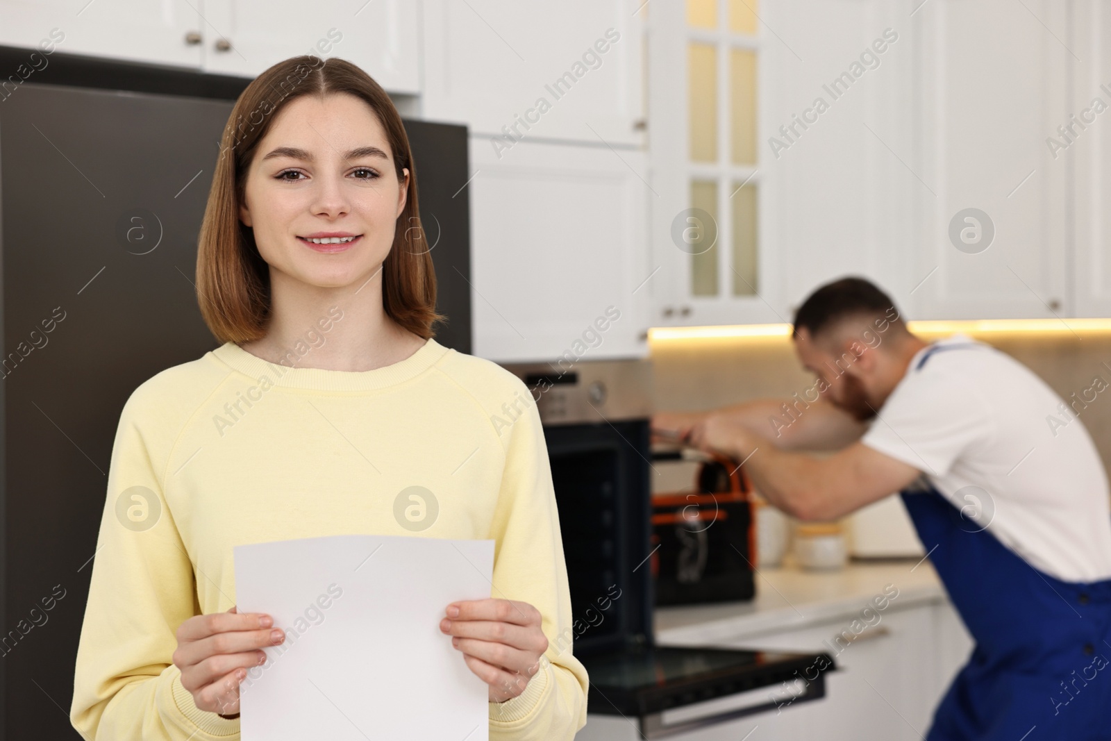 Photo of Smiling woman with sheet of paper and repairman fixing oven in kitchen, selective focus