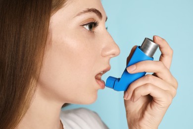 Young woman using asthma inhaler on light blue background, closeup