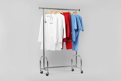 Photo of Different medical workers' uniforms and stethoscope on clothing rack against white background