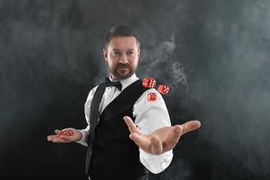 Photo of Professional croupier holding dice and casino chips on black background with smoke