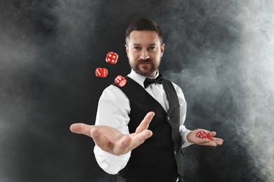 Professional croupier holding dice and casino chips on black background with smoke