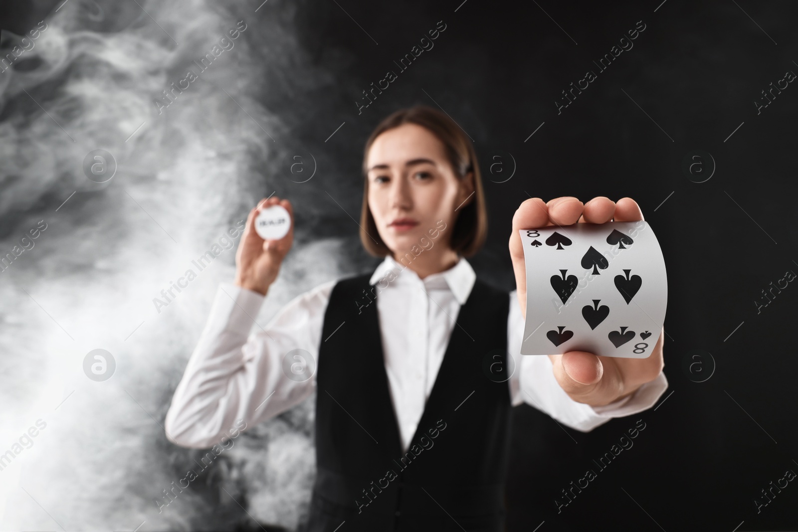 Photo of Croupier holding cards and dealer button on black background with smoke, selective focus