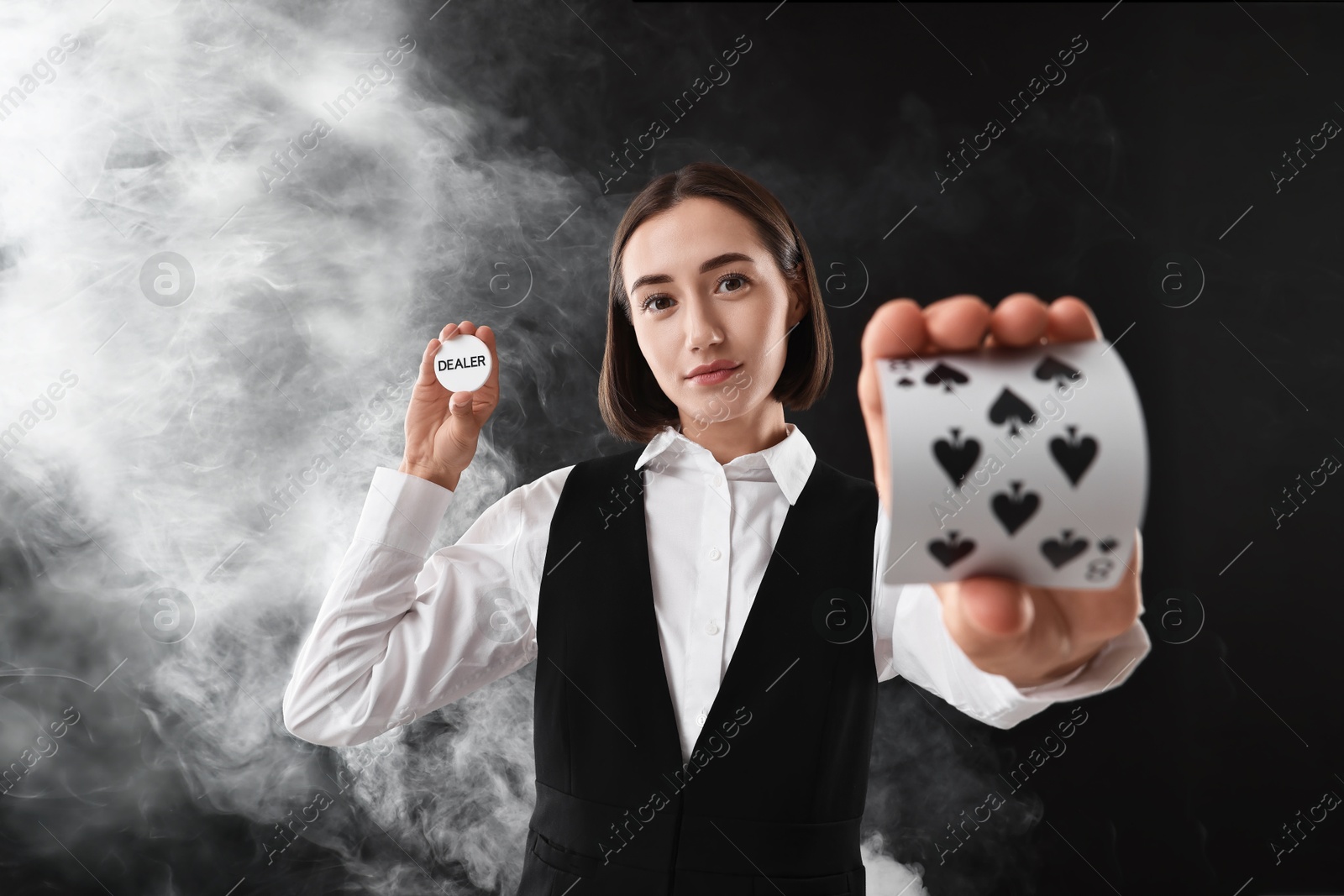 Photo of Croupier holding cards and dealer button on black background with smoke, selective focus