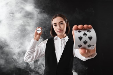 Photo of Croupier holding cards and dealer button on black background with smoke, selective focus