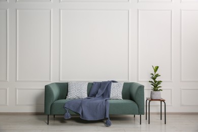 Stylish sofa with cushions, blanket and plant on side table near white wall