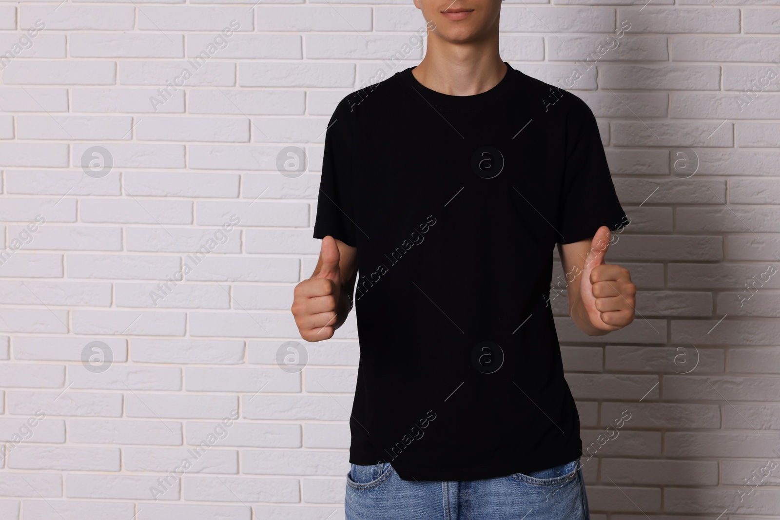 Photo of Teenage boy wearing black t-shirt and showing thumbs up near white brick wall, closeup. Space for text