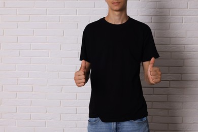 Photo of Teenage boy wearing black t-shirt and showing thumbs up near white brick wall, closeup. Space for text