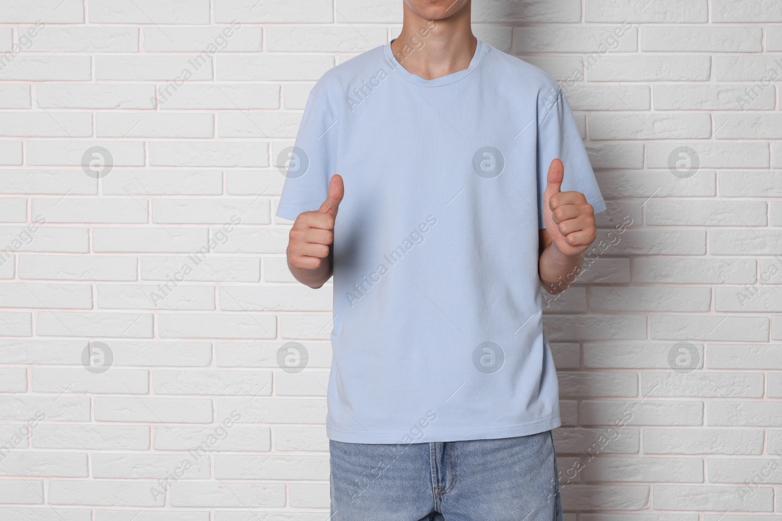 Photo of Teenage boy wearing light blue t-shirt and showing thumbs up near white brick wall, closeup. Space for text