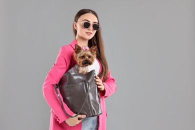 Photo of Woman in sunglasses carrying cute Yorkshire Terrier dog in bag on grey background, space for text