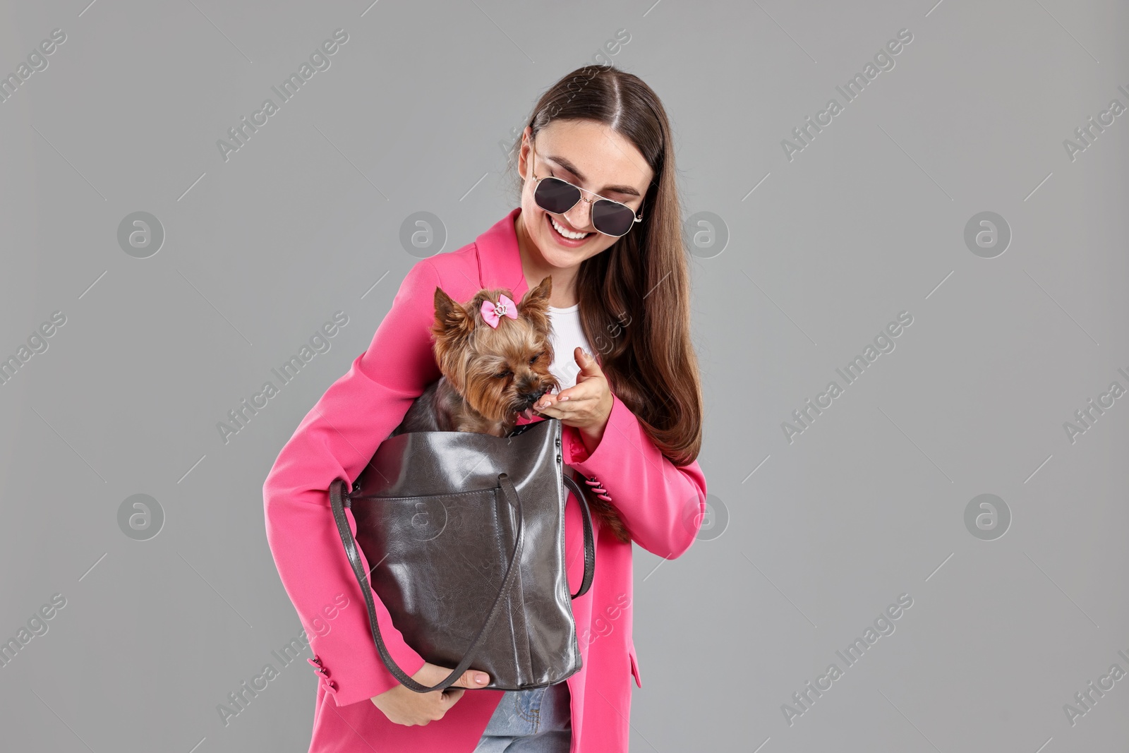 Photo of Woman in sunglasses carrying cute Yorkshire Terrier dog in bag on grey background