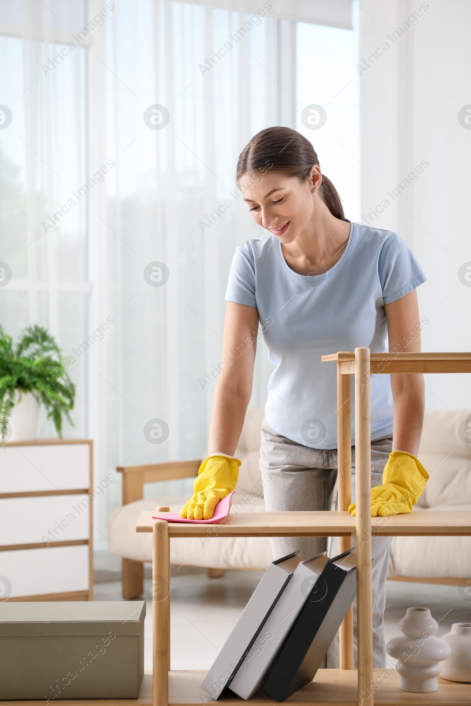 Photo of Young woman cleaning wooden shelf with rag at home