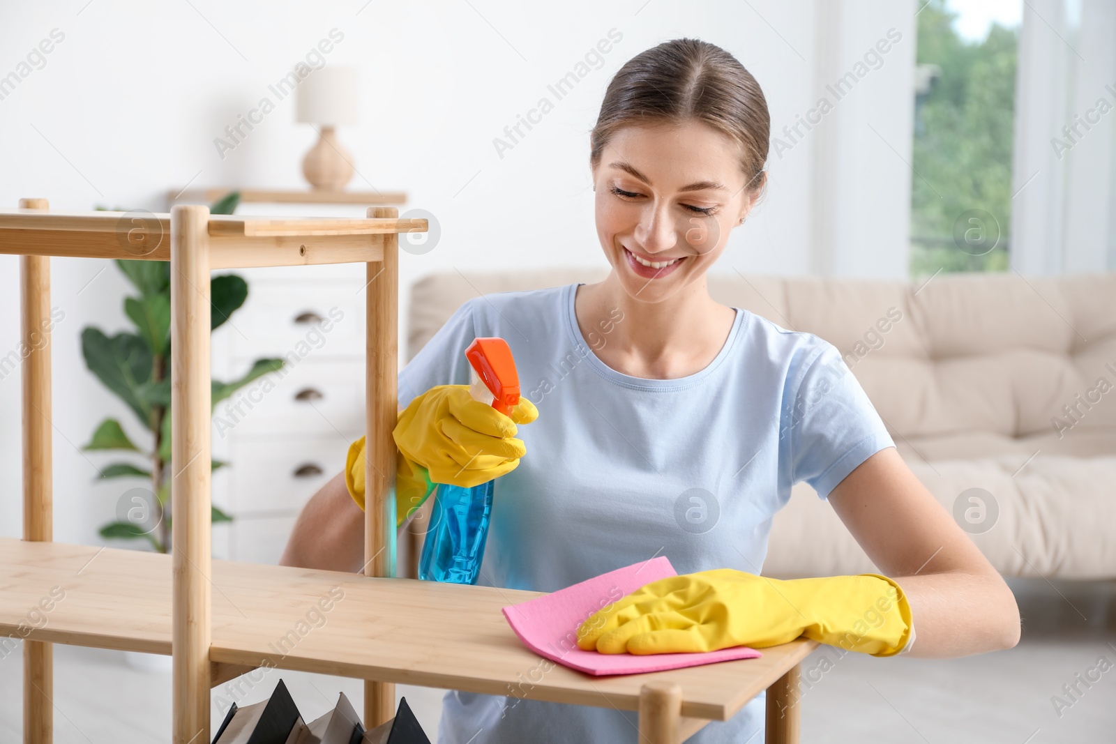 Photo of Young woman cleaning wooden shelf with rag and spray at home