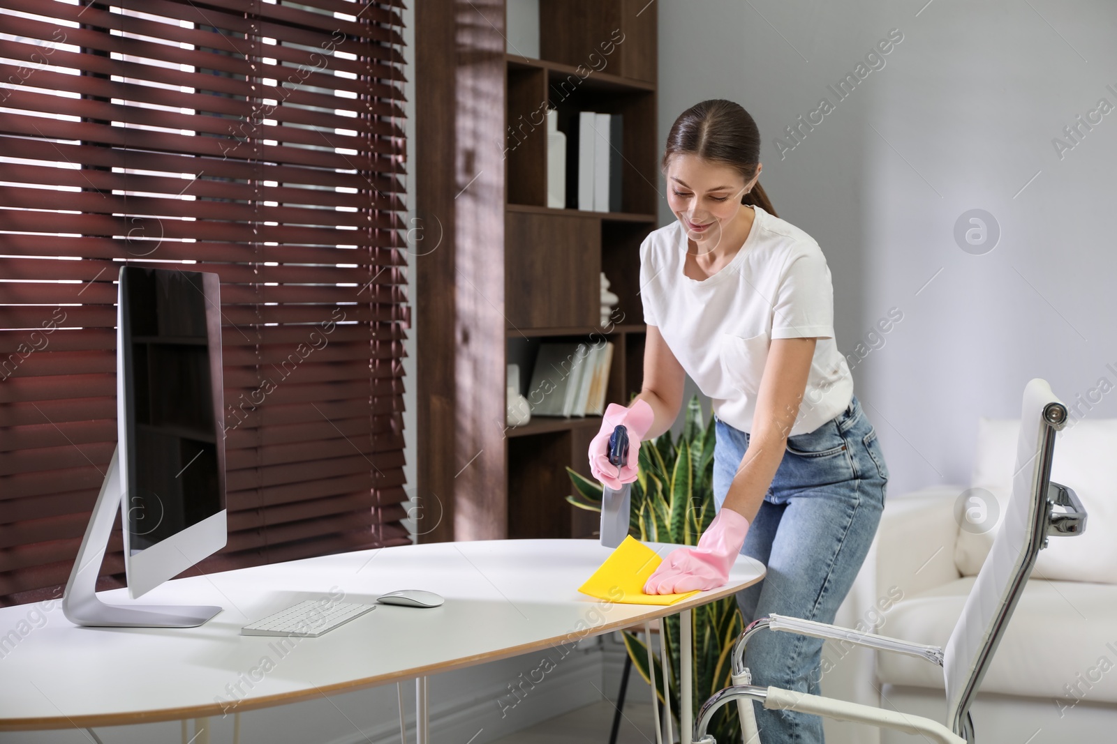 Photo of Young woman cleaning table with rag and spray in office