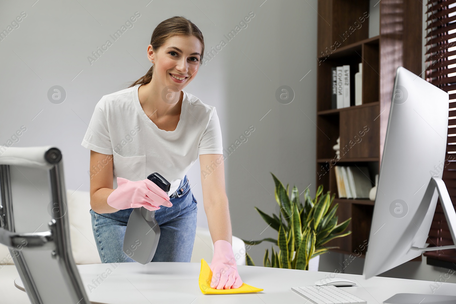 Photo of Young woman cleaning table with rag and spray in office