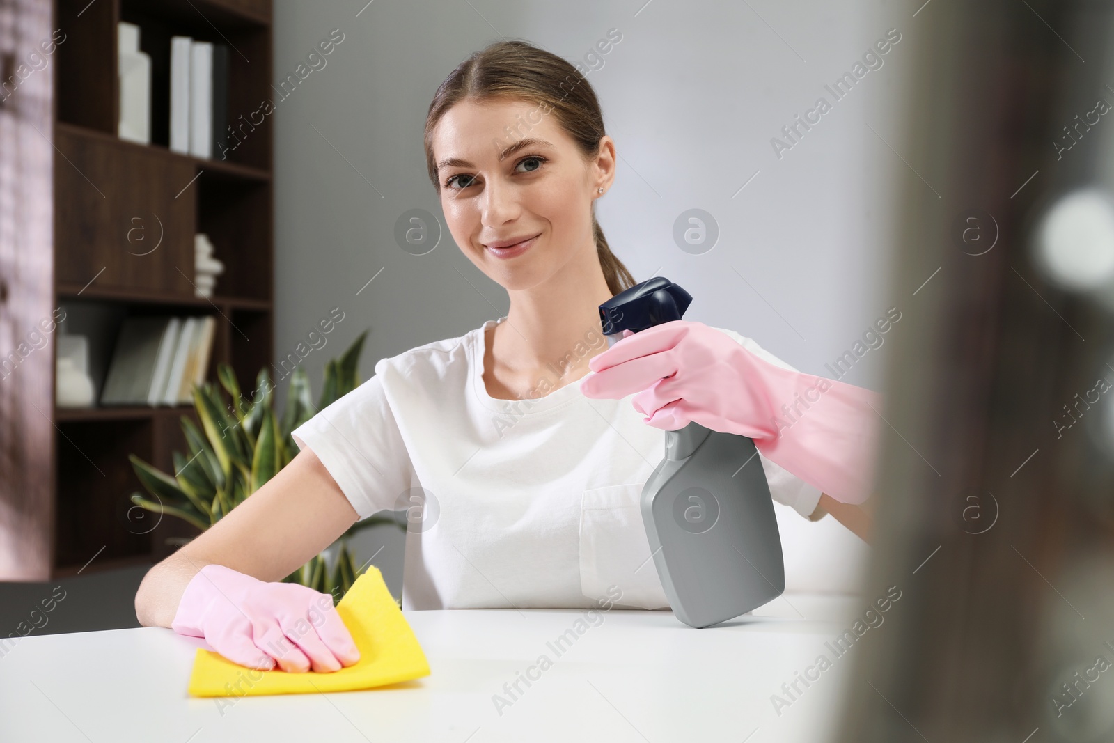 Photo of Young woman cleaning table with rag and spray in office